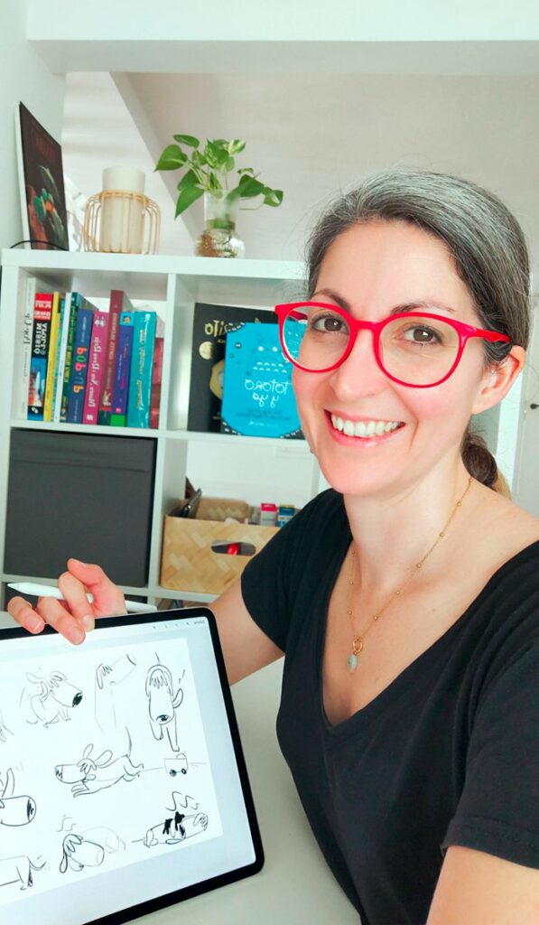 mireia_crea_crop Mireia Serra Poch somrient mentre mostra esbossos de personatges canins en una tauleta digital, amb prestatgeria de llibres de fons